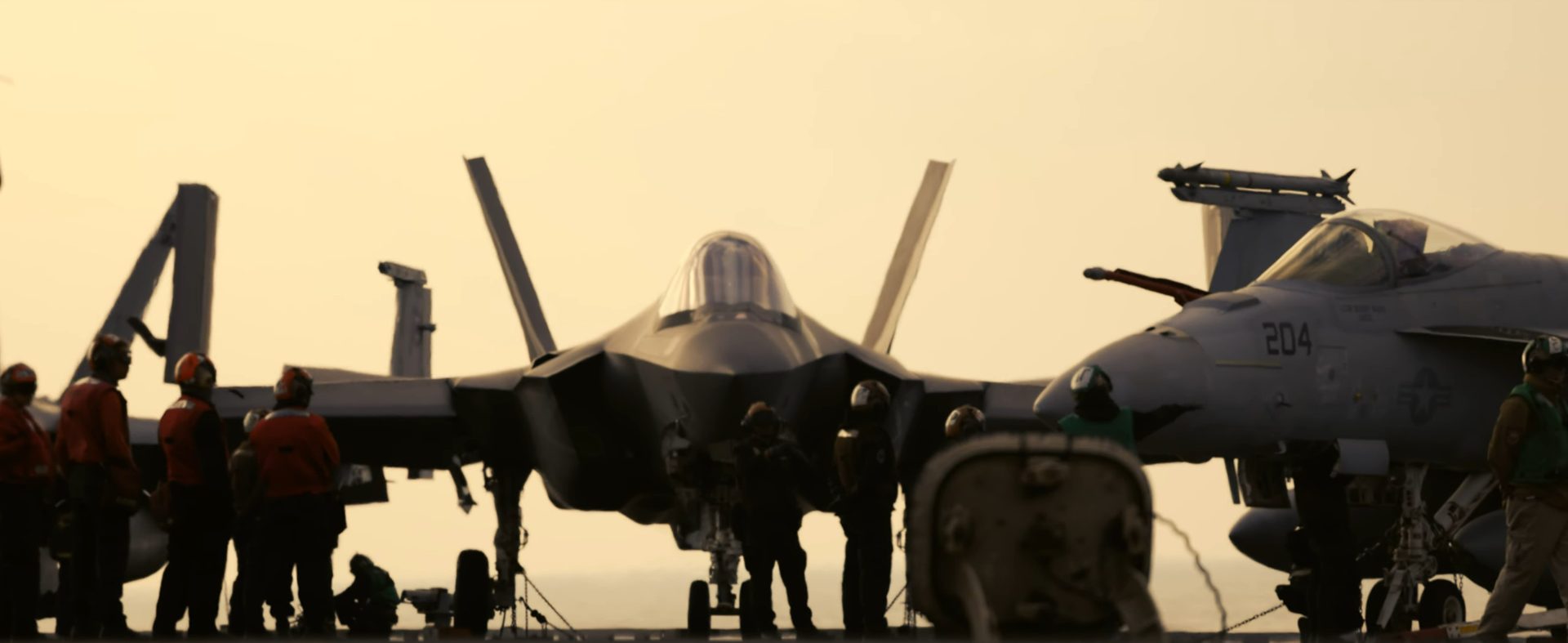 Deck crew around an F-35C and a Hornet on the carrier deck in haze